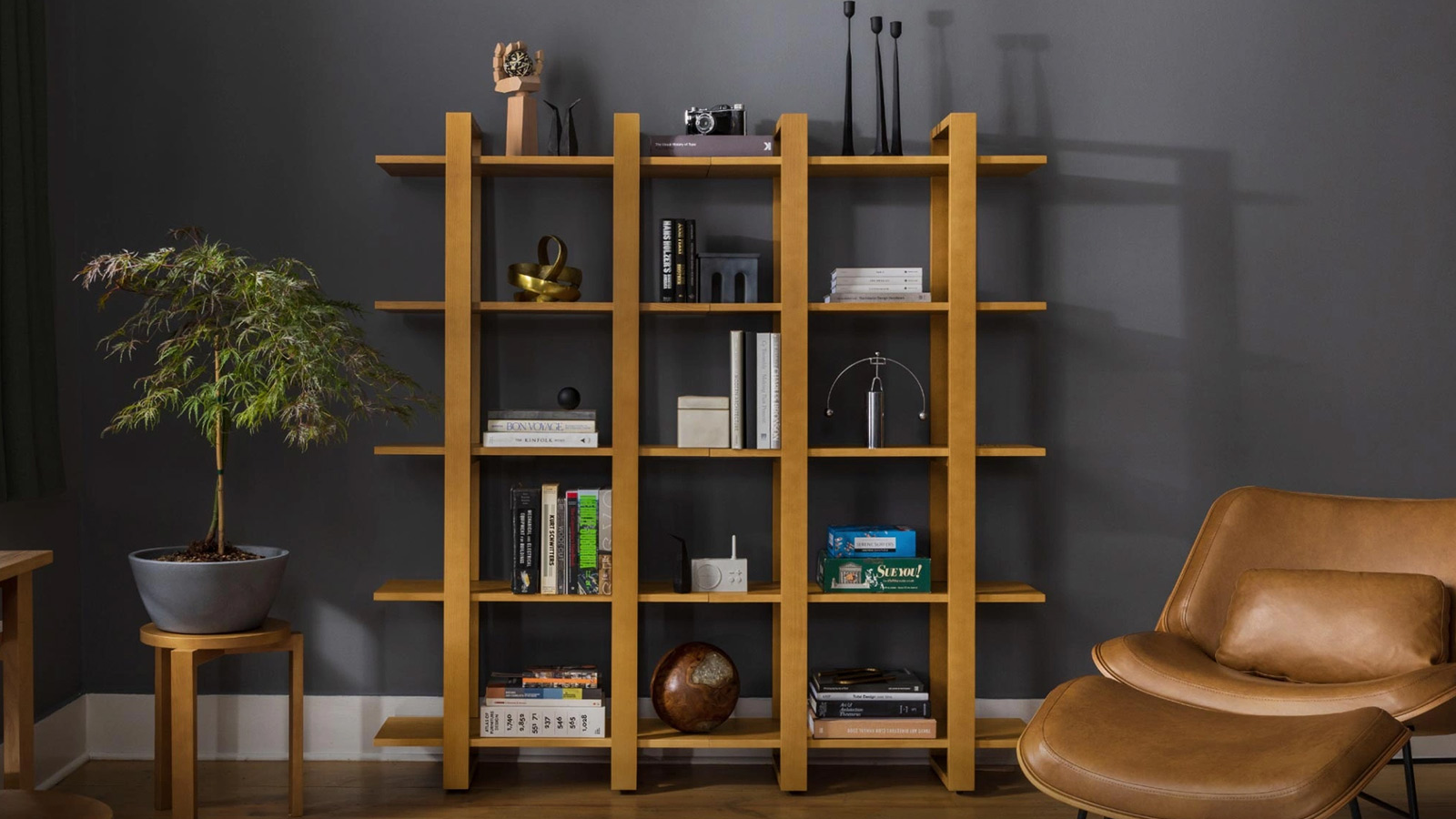 The image features a contemporary living space with a dark gray wall. Centered in the image is a large, light wooden bookshelf filled with various objects. The bookshelf houses an assortment of items including books, decorative sculptures, a small plant, a camera, a brass bowl, a globe, and some electronic devices. To the left, there is a small wooden table upon which a potted Japanese maple bonsai tree is placed. To the right, there's a modern leather lounge chair with a matching ottoman, both in a caramel brown color.