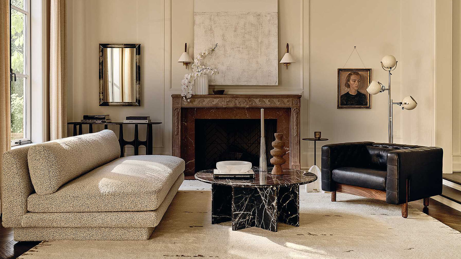 This image presents a well-appointed living room with a classic and sophisticated interior. The room features a high ceiling and paneled walls painted in a light cream color, complemented by floor-length drapes framing a window that lets in natural light. The furnishings include a plush, textured fabric chaise lounge and a dark leather armchair, adding a contrast of textures. A circular marble coffee table with dark veins stands at the center of the room on a large, light-colored area rug. To the right, a metal floor lamp with three globular shades adds a modern touch next to a framed portrait of a woman hanging on the wall. In the center, above an ornate marble fireplace, hangs a large abstract canvas flanked by two classic wall sconces. On the left, a sleek console table holds books, and a rectangular mirror above reflects part of the room.