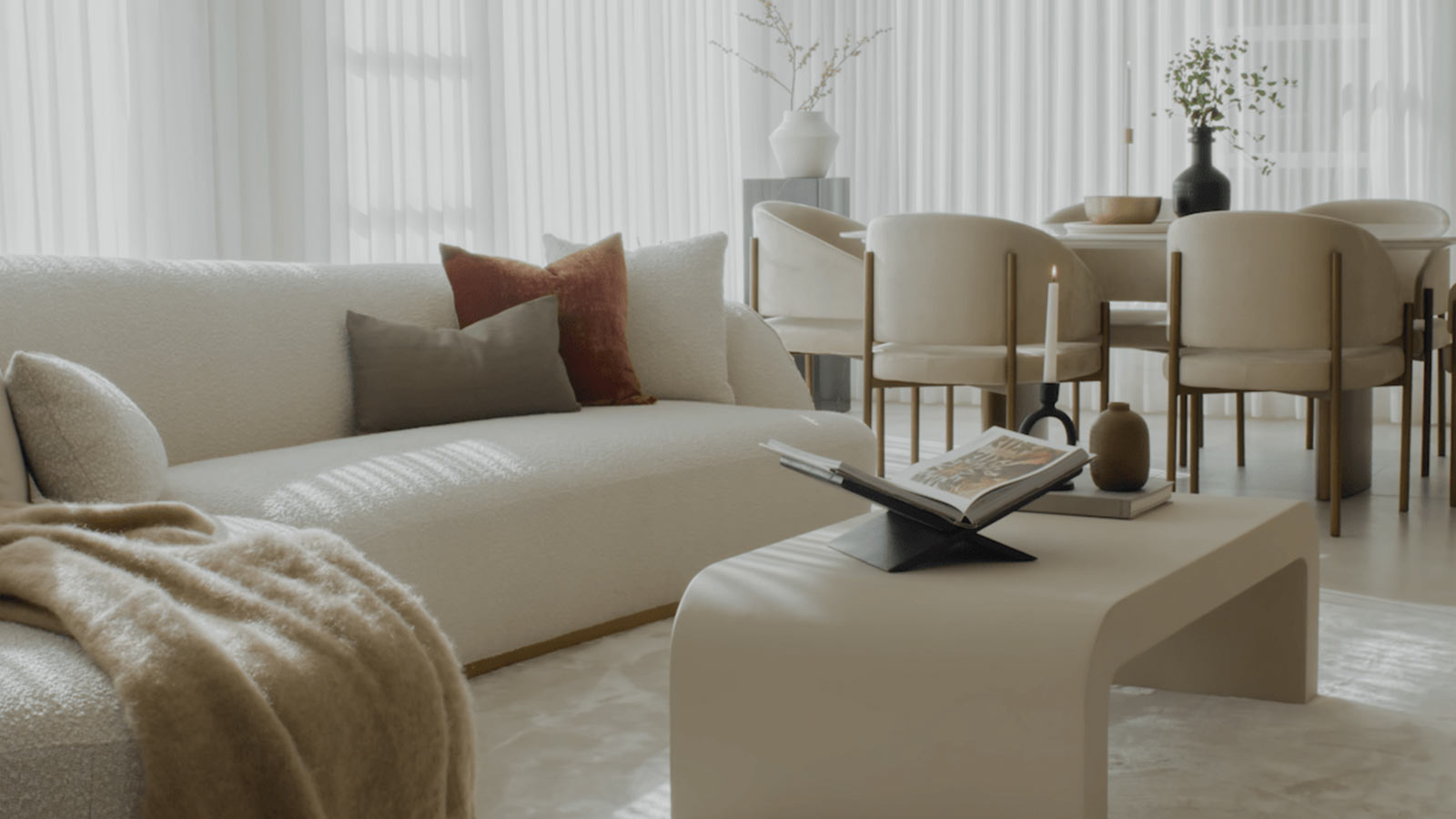 This image captures a serene and minimalist living space bathed in natural light. In the foreground, a large, plush sofa in a textured off-white fabric is adorned with decorative pillows in muted earth tones. A cozy beige throw blanket is draped over one end of the sofa. A sleek, modern coffee table in front of the sofa holds a neatly stacked collection of hardcover books, one open as if recently browsed through. In the background, a dining area is visible, featuring a round table surrounded by elegant upholstered chairs, and a simple yet stylish vase with sprigs of greenery atop the table. Floor-to-ceiling sheer curtains filter the sunlight, creating a soft and tranquil atmosphere. The color palette is neutral, with whites and creams predominating, accented by the natural colors of the books and foliage, which add a touch of warmth to the space. The overall effect is one of calm sophistication and understated luxury.