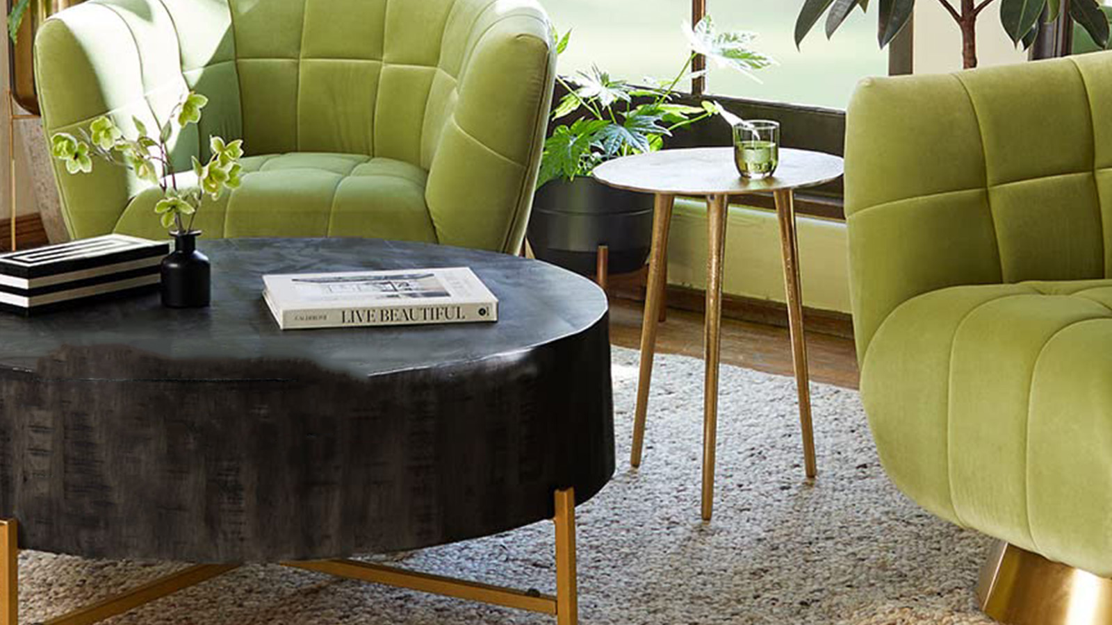 This is an interior scene featuring modern furniture. In the foreground is a coffee table with a dark, glossy surface, sitting on golden legs. On the table are a striped black-and-white book, a small black vase with green flowers, and a large book titled "LIVE BEAUTIFUL". To the right, there's a small side table with a marble top and golden legs, holding a clear glass with a greenish liquid. Two plush, lime green armchairs with tufted backrests flank the tables, their velvety fabric catching the light.
