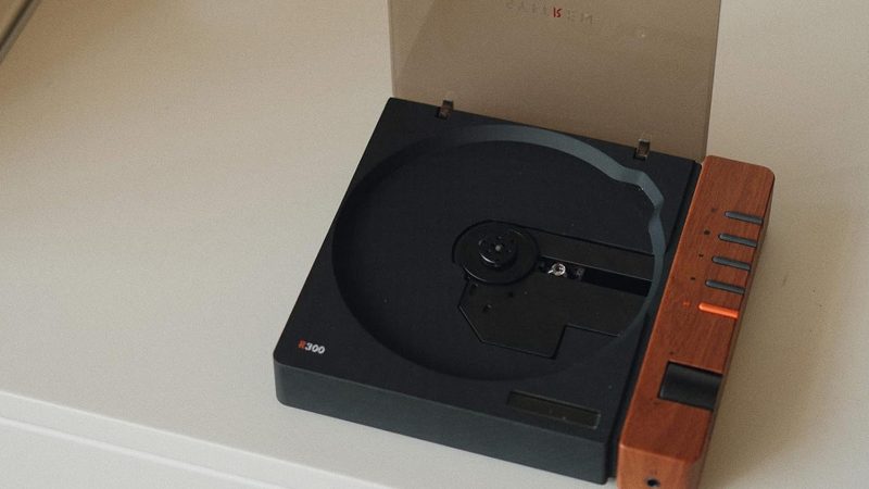 The Syitren R300 CD Player boasts a matte black design with a wood-paneled control zone, a transparent disc lid, a tactile volume knob, Bluetooth, AUX, optical output, and a USB-C power port.