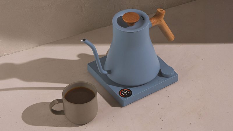 Fellow Stagg EKG electric kettle in matte blue with a wood handle, shown with a ceramic mug—an example of design-forward everyday accessories for coffee rituals.