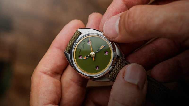 The Singer Caballero watch with stainless steel C-shaped case, green, black, or blue lacquered dial, inset rubies, and signature orange-lumed hands on a textile or leather strap.