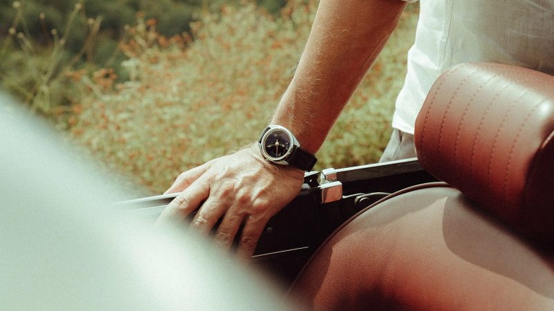 The Singer Caballero watch with stainless steel C-shaped case, green, black, or blue lacquered dial, inset rubies, and signature orange-lumed hands on a textile or leather strap.