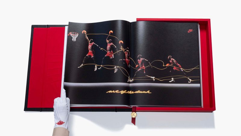A deluxe edition book titled ‘Air Jordan: The Ultimate Collection’ by Assouline, encased in an ornate black leather clamshell box embossed with Baroque motifs, with a red ribbon and gold medallion bookmark visible.