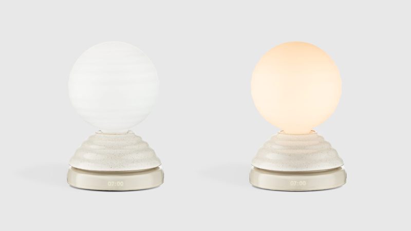 Tala WAKE Sleep Light by Heatherwick Studio emits a soft sunrise-inspired glow beside a bed.