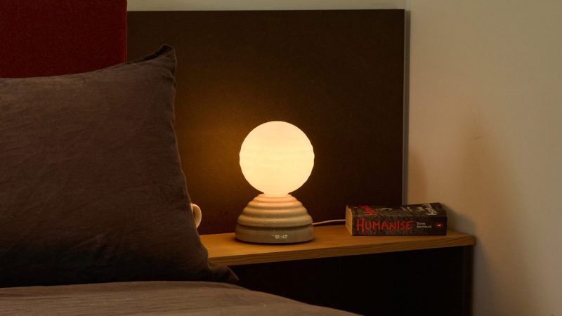 Tala WAKE Sleep Light by Heatherwick Studio emits a soft sunrise-inspired glow beside a bed.