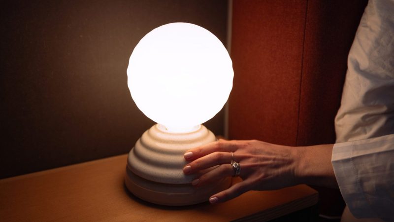 Tala WAKE Sleep Light by Heatherwick Studio emits a soft sunrise-inspired glow beside a bed.