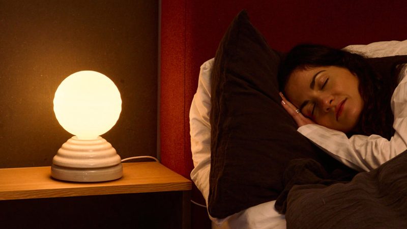 Tala WAKE Sleep Light by Heatherwick Studio emits a soft sunrise-inspired glow beside a bed.