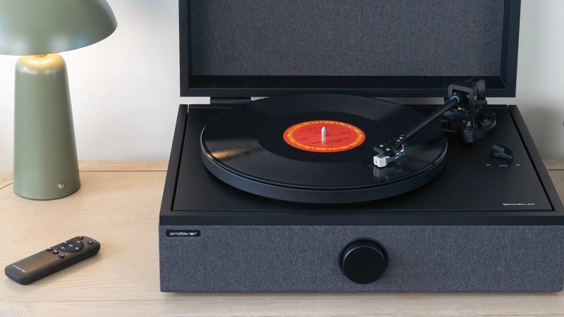 High-end all-in-one record player system by Andover Audio: a sleek black turntable with built-in amp and speakers, hard-cover lid open, vinyl spinning, minimalist aesthetic.
