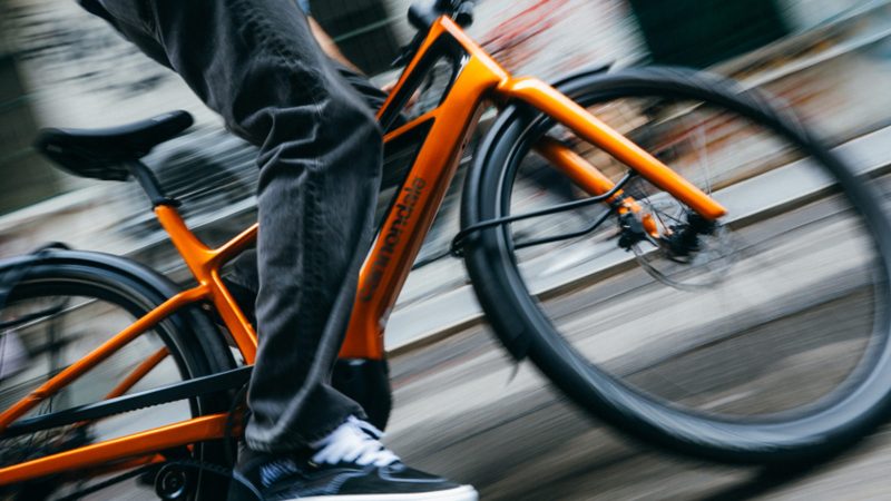 The Cannondale FlyingV carbon e-bike with V-frame design, carbon fenders, and belt-drive system displayed in an urban setting.