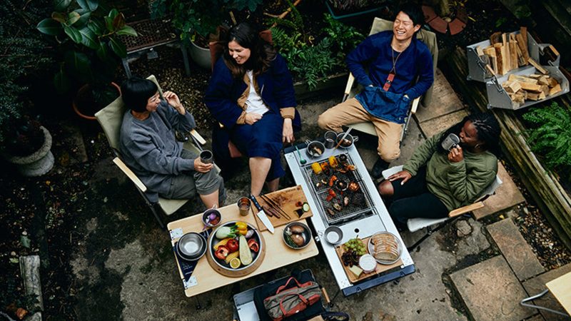 Snow Peak Iron Grill camp kitchen and table setup outdoors features modular stainless steel cooking units and a wooden tabletop for outdoor dining.