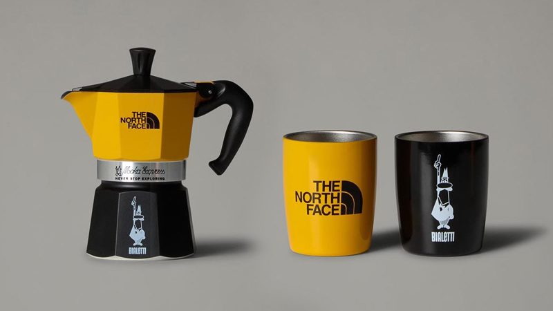 The North Face x Bialetti Moka Express Set features a co-branded stovetop espresso maker and camping mug in an outdoor alpine setting, designed for trail-side coffee brewing.