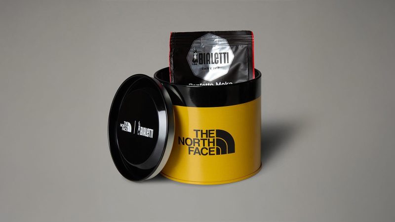 The North Face x Bialetti Moka Express Set features a co-branded stovetop espresso maker and camping mug in an outdoor alpine setting, designed for trail-side coffee brewing.