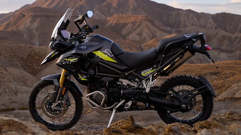 Triumph Tiger 1200 Desert Edition off-road motorcycle navigating sand dunes, matte sand-colored body, rugged knobby tires, desert backdrop.