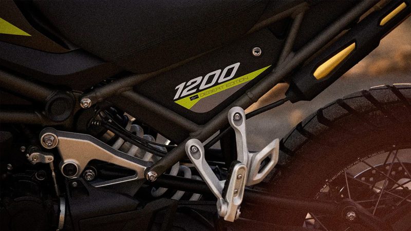 Triumph Tiger 1200 Desert Edition off-road motorcycle navigating sand dunes, matte sand-colored body, rugged knobby tires, desert backdrop.