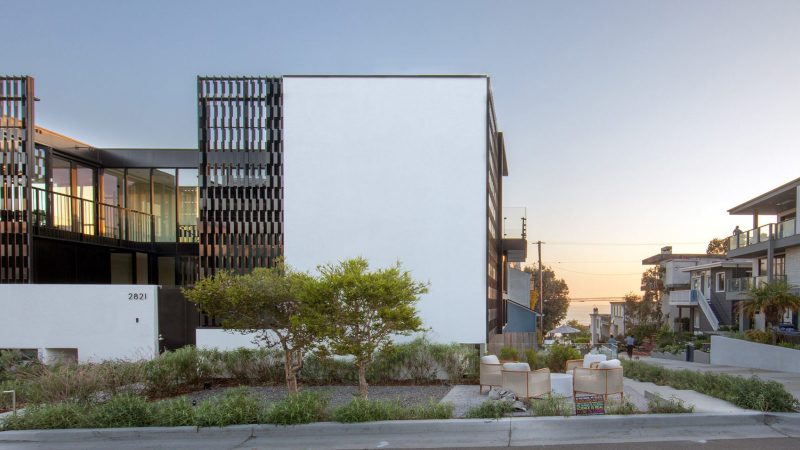 The Alma Switch House in Manhattan Beach features a modern white stucco facade, a shimmering silver aluminum privacy screen, and an open-air central courtyard.